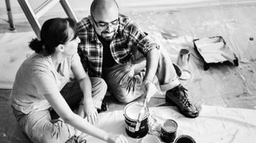 Photo of couple sitting on floor with paint buckets and brushes in hand