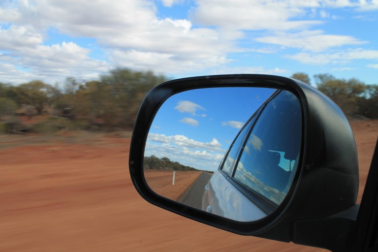 The view of a red dirt road from a car rearview mirror.