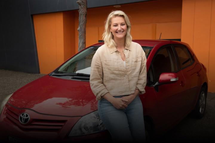 A blonde happy looking woman smiling and leaning against her car.